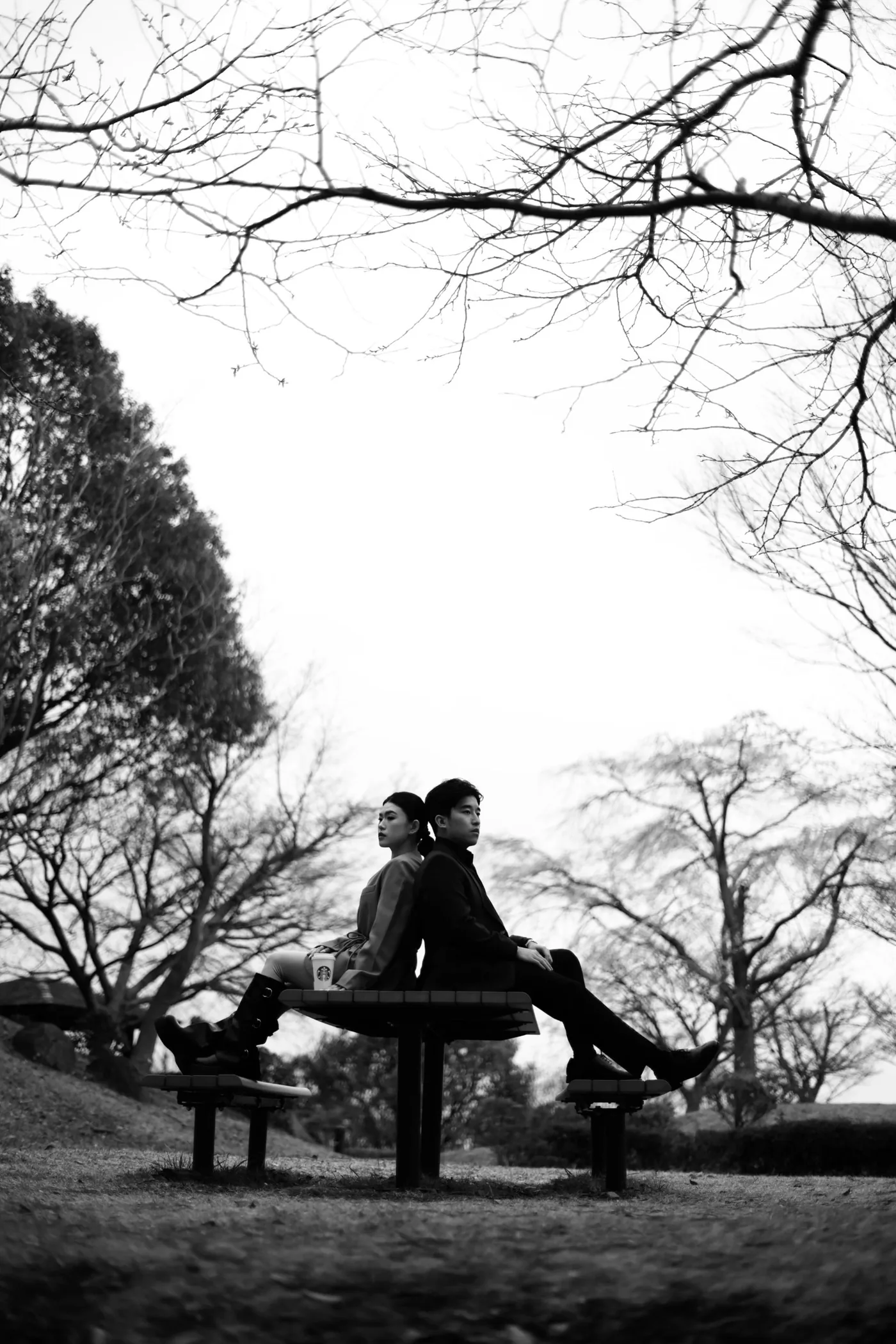 Louis and Anezka, creative directors of Caspia Fleur, sitting back to back on a park bench beneath bare winter branches, in black and white