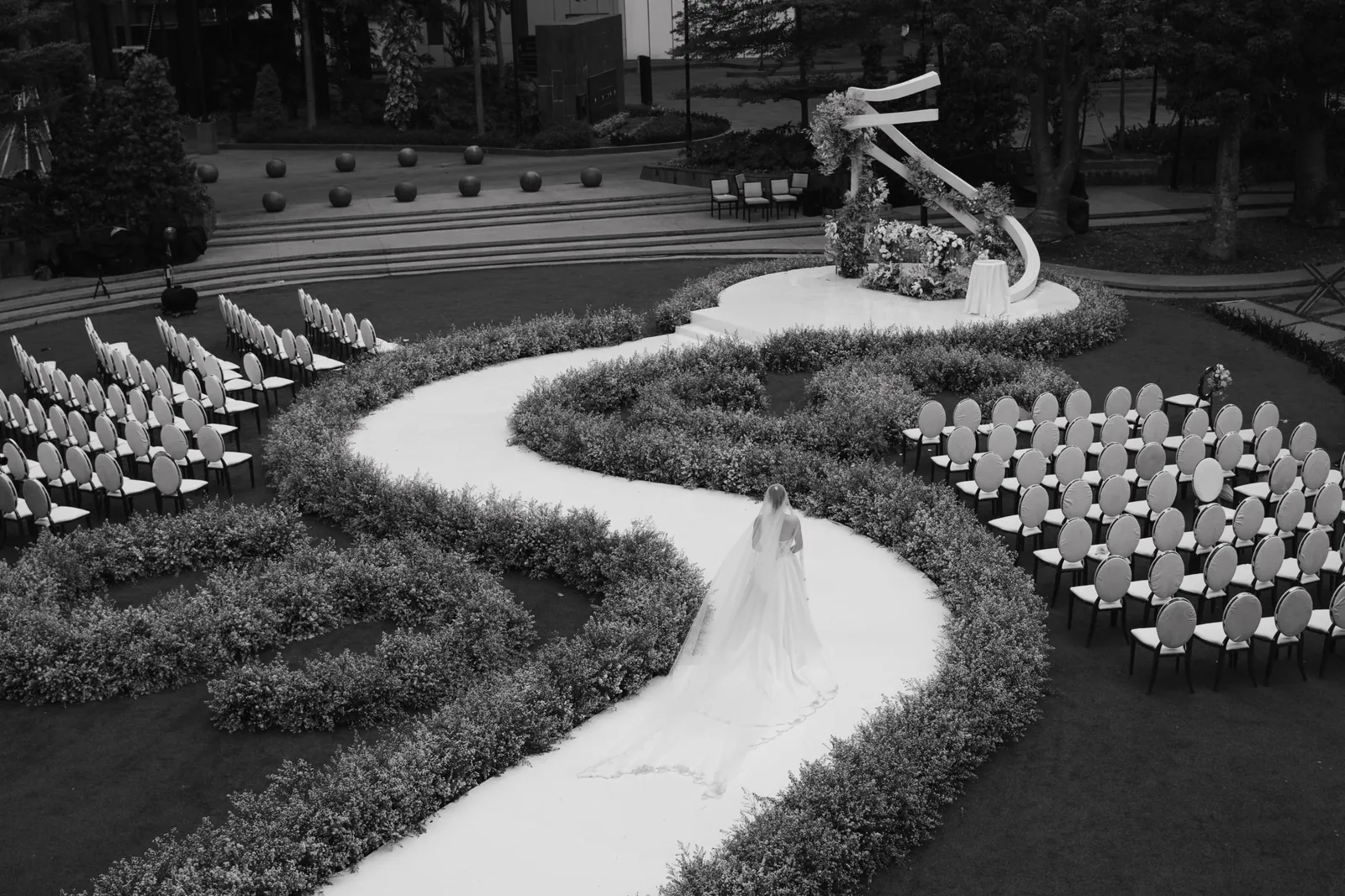 A bride walks a winding white aisle through low floral hedges toward a sculptural ribbon arch, in black and white
