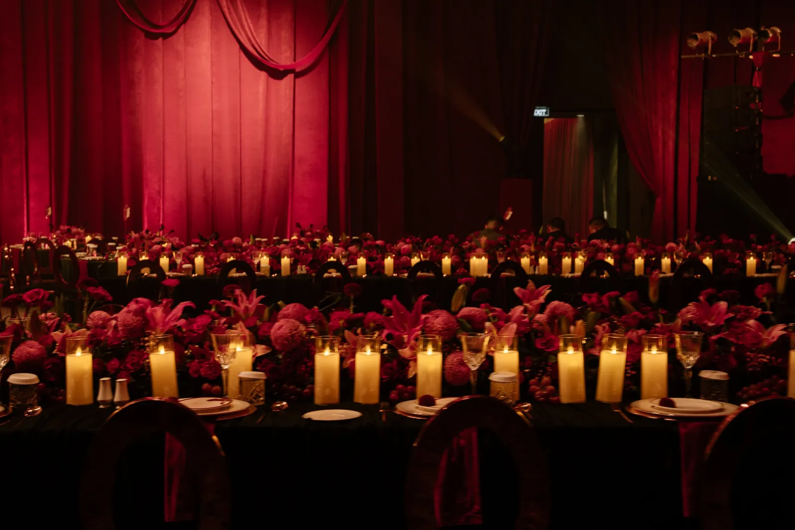 Long banquet tables lined with pillar candles and pink and red florals before a crimson curtain