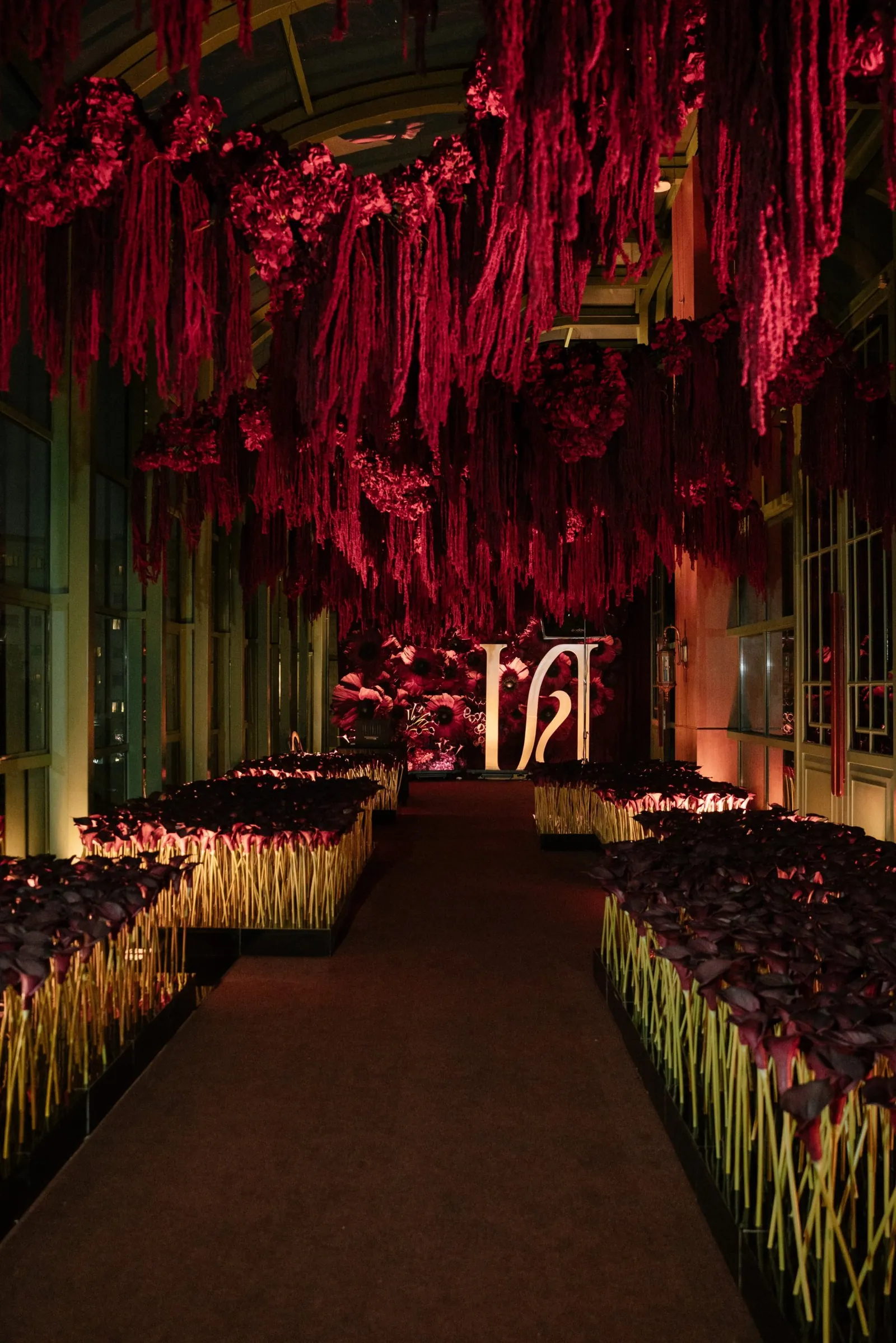 A corridor hung with cascading crimson amaranthus above beds of dark calla lilies, leading to a glowing monogram