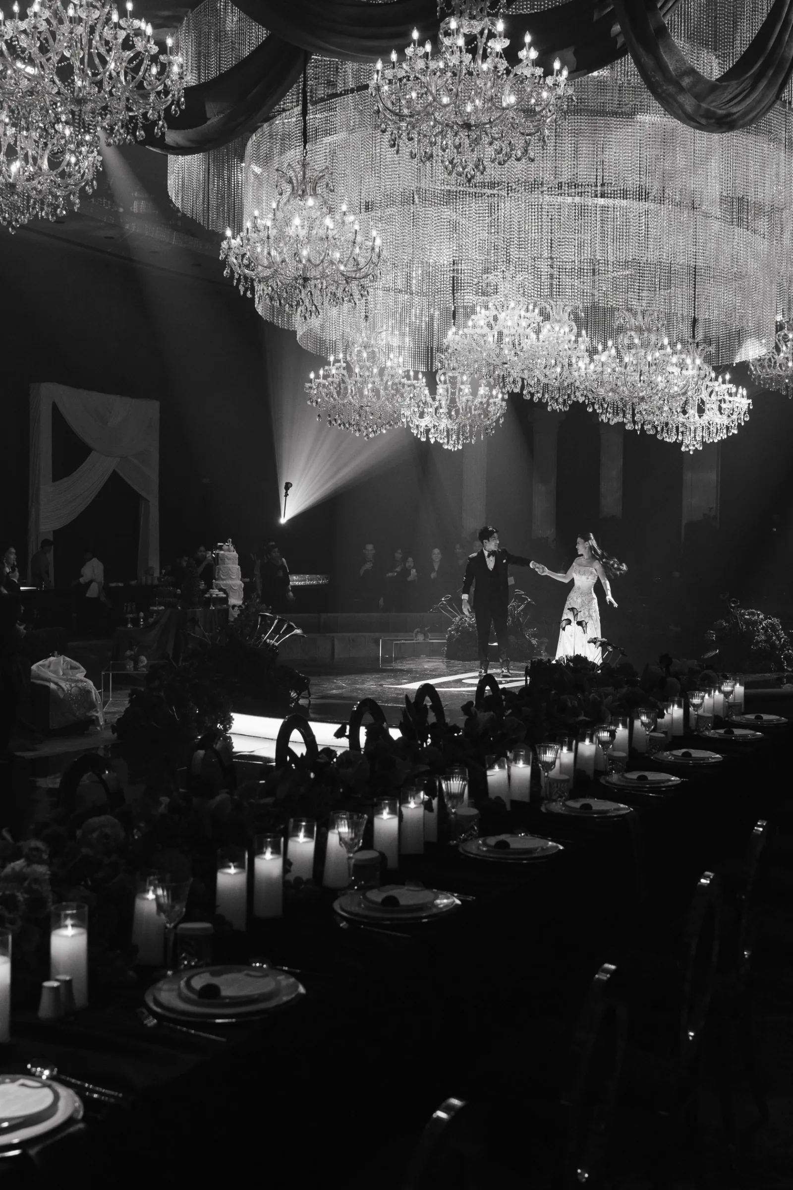 A couple shares their first dance beneath tiered crystal chandeliers, past candlelit banquet tables, in black and white