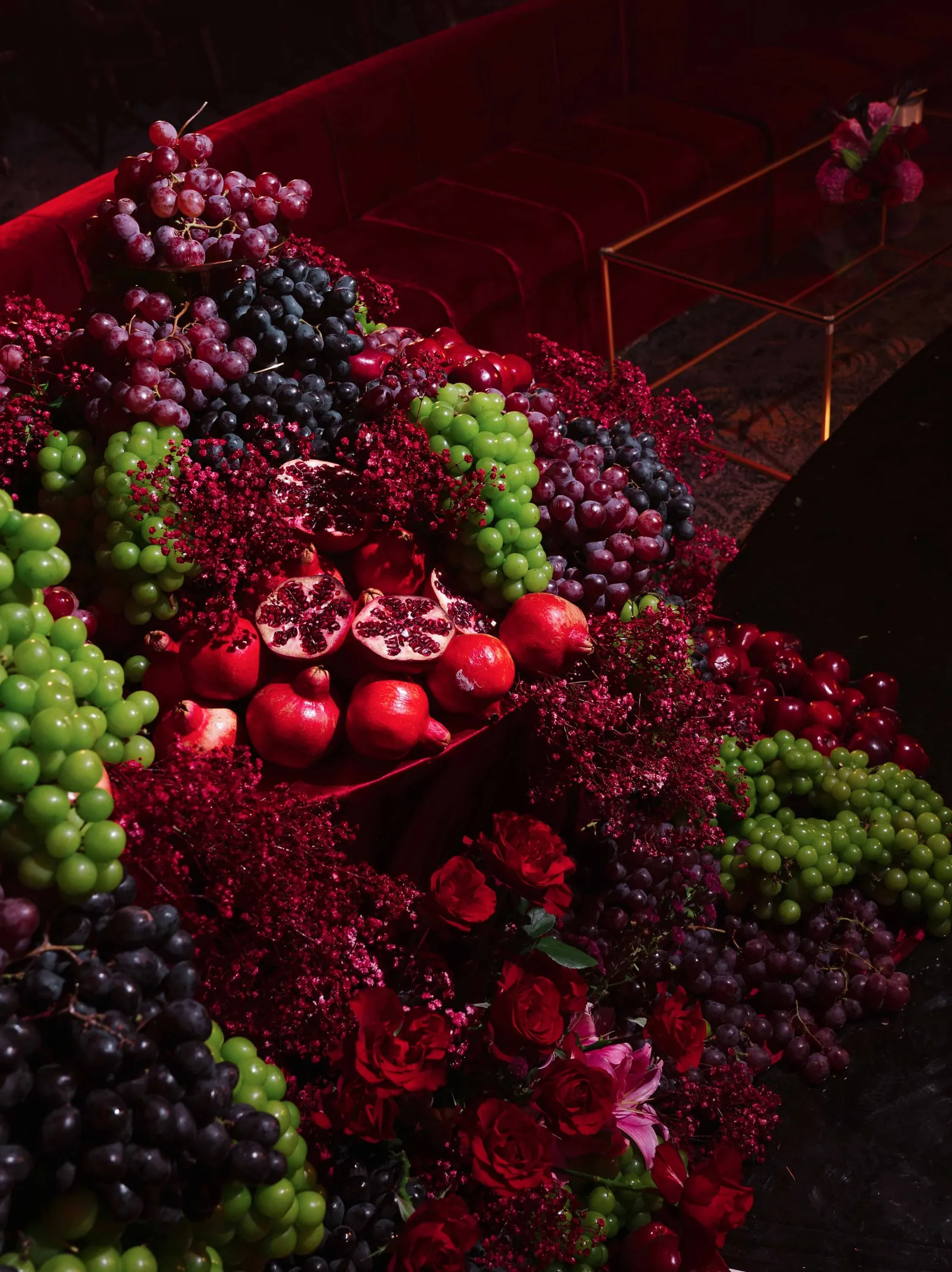 A cascade of grapes, halved pomegranates, red roses and crimson gypsophila beside a velvet sofa
