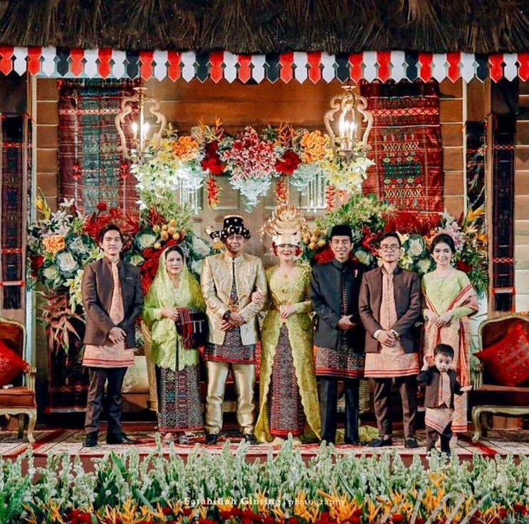 The bridal party and families in traditional dress on a decorated wedding stage with Mandailing textiles and florals
