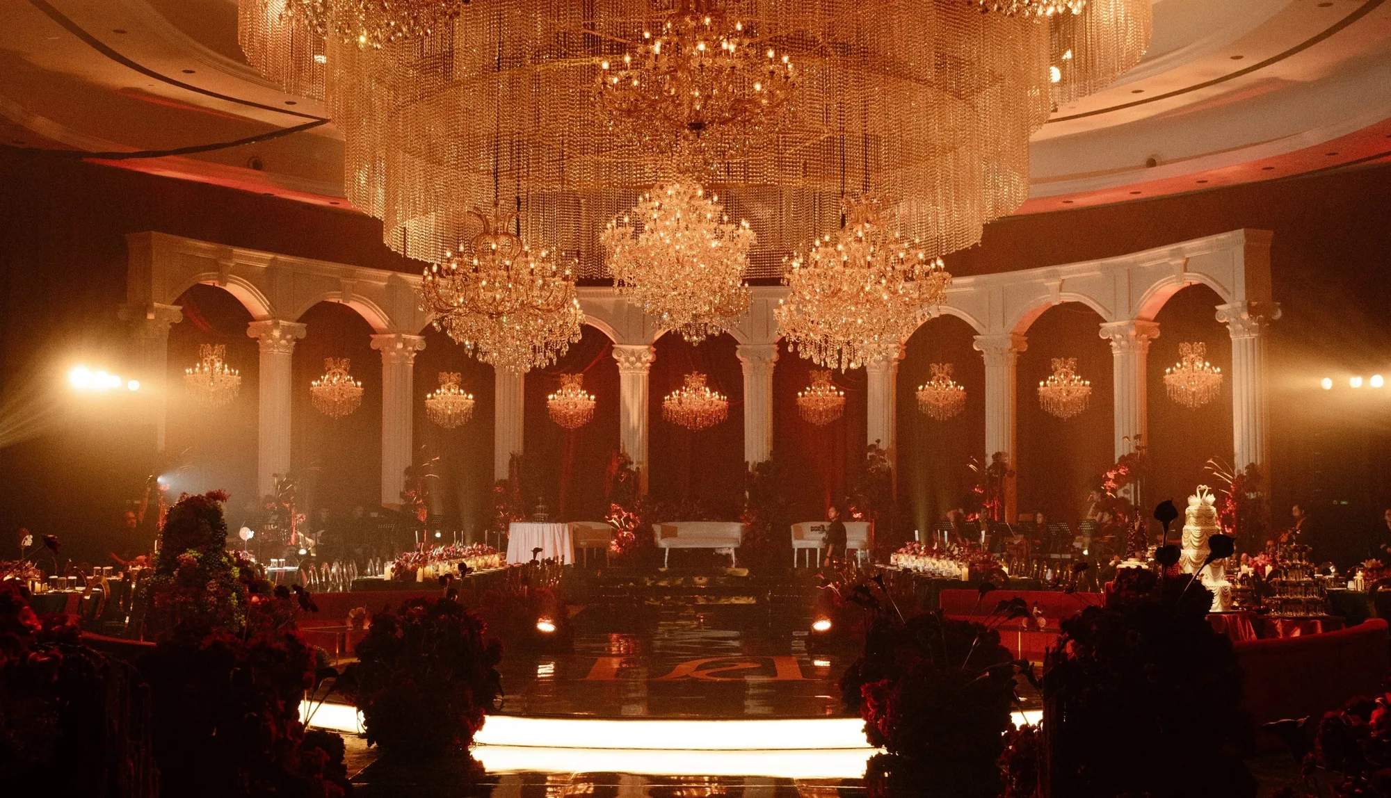 Ballroom under tiered crystal chandeliers and crimson drapes, with an ivory colonnade, deep red florals and a mirrored aisle leading to a glowing stage